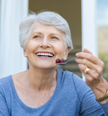 Cheerful senior woman holding red grapes in spoon and make a beautiful white smile. Smiling old woman looking away while eating fresh fruits for breakfast. Mature woman enjoying old age.