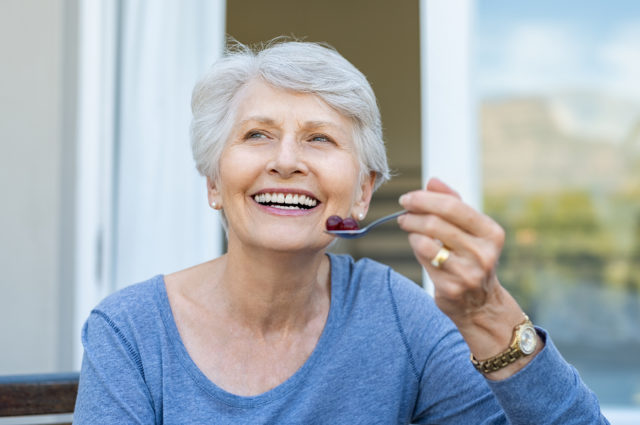 Cheerful senior woman holding red grapes in spoon and make a bea